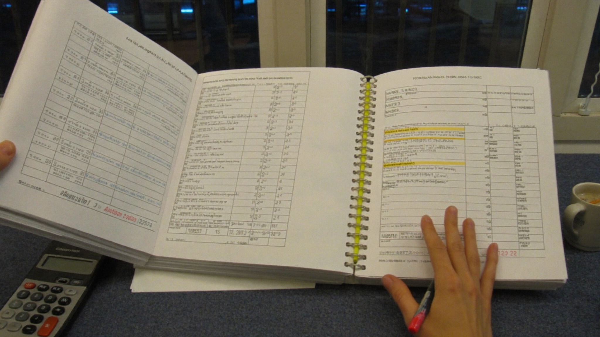 A payroll register open on a desk at night, rows highlighted in yellow, a calculator and a cold mug beside it.