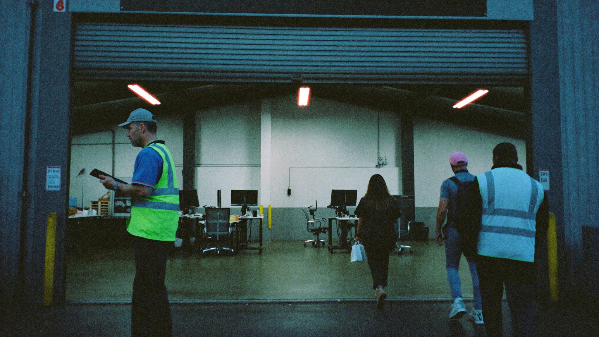 A supervisor in a hi-vis vest with a clipboard at an open roller door before dawn, workers walking in past him, empty desks and chairs visible inside.